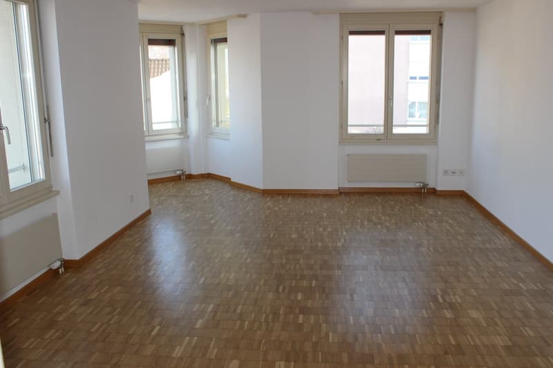 Empty room with wooden floors, white walls, three windows, and radiators.