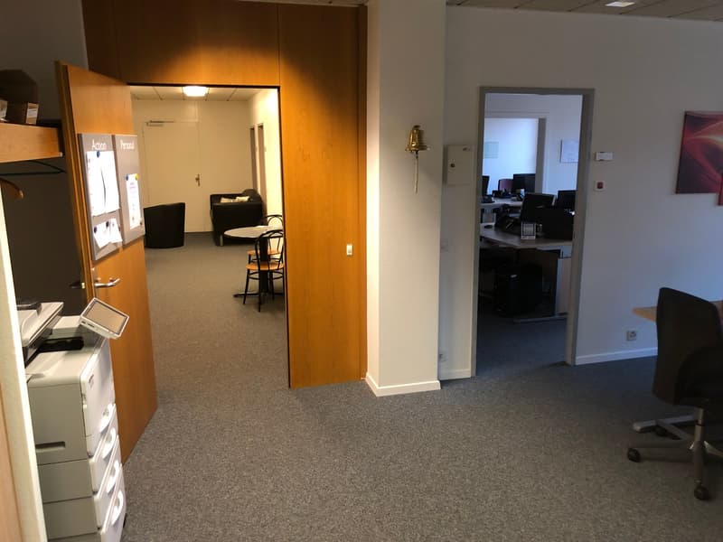 The image shows an office space with a carpeted floor, wooden doors, and various office equipment such as a printer and computer monitors. The room appears to have a waiting area with a chair and a small table.
