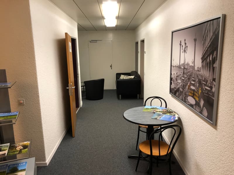The image shows an office space with a round table, two black chairs, and a large framed artwork on the wall. The room has a carpeted floor, white walls, and recessed lighting. There is a door visible in the background.