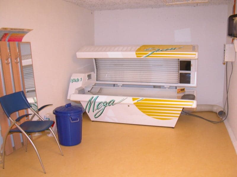 A tanning bed is in the corner, blue chair and blue trash can nearby, mirror on the wall, wooden cabinet, power outlet on the wall