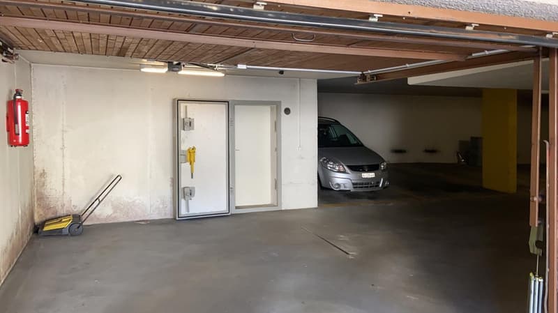 A clean concrete floor, a silver car parked, a white door, fire extinguisher mounted on the wall