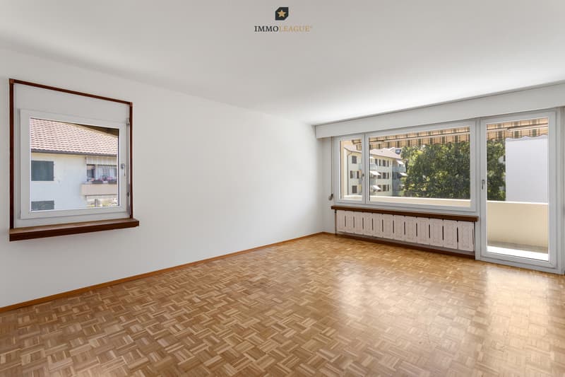 Grosszügiges Wohnzimmer, das mit warmem Parkett und grossen Fenstern viel Licht ins Haus holt und direkten Zugang zum sonnigen Südbalkon bietet.