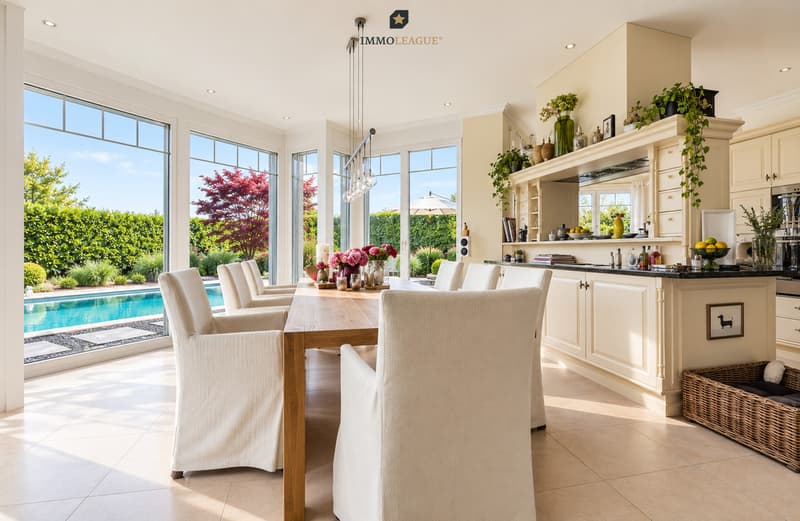 Eine offene Küche mit Essbereich und großen Fenstern bietet den Blick auf ein modernes Haus mit Blick auf den Pool / an open kitchen and dining area with large windows opens up to a modern home with