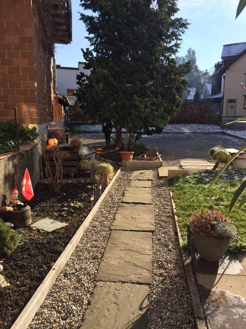 Garden with plants, gravel path, flag, outdoor gnome, and a person standing by a tree