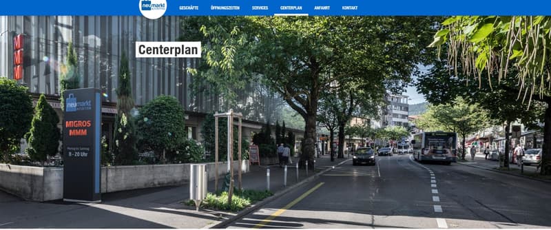 The image shows a street view of a commercial area with various shops and businesses. There are trees lining the street, and the buildings have a mix of modern and traditional architectural styles. The street appears to be a busy commercial district with