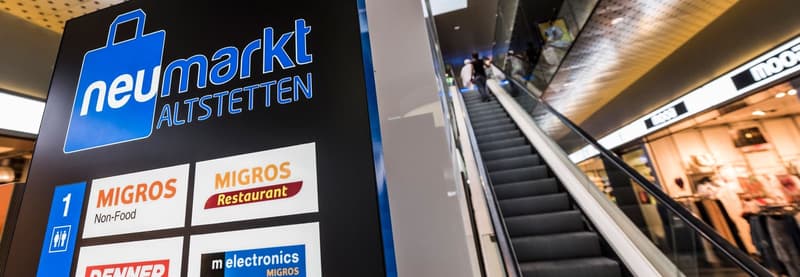 The image shows a large sign for 'Neumarkt Altstetten', which appears to be a shopping mall or commercial center. The sign lists the various stores and amenities available, including Migros (a non-food store), Migros Restaurant, and m electronics Migros.