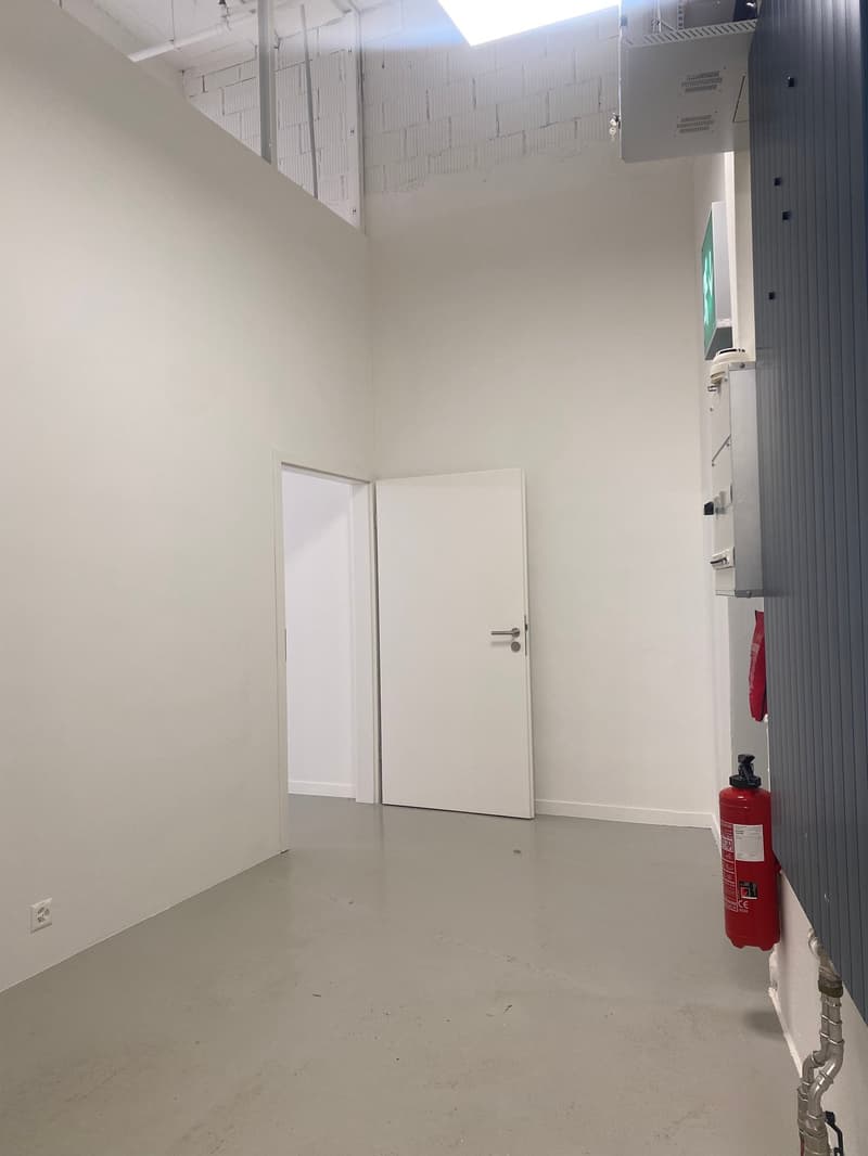 The image shows a small, empty room with white walls and a concrete floor. There is a white door on the right side of the room, and a fire extinguisher mounted on the wall. The ceiling appears to be made of a grid of suspended panels.