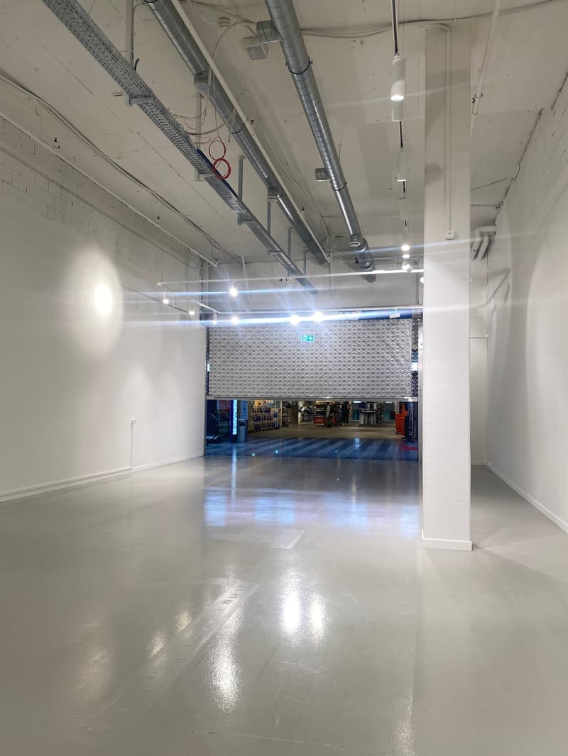 The image shows a large, open commercial space with a high ceiling, exposed ductwork, and a polished concrete floor. The space appears to be a warehouse or industrial-style commercial area with a metal rolling door visible in the background.