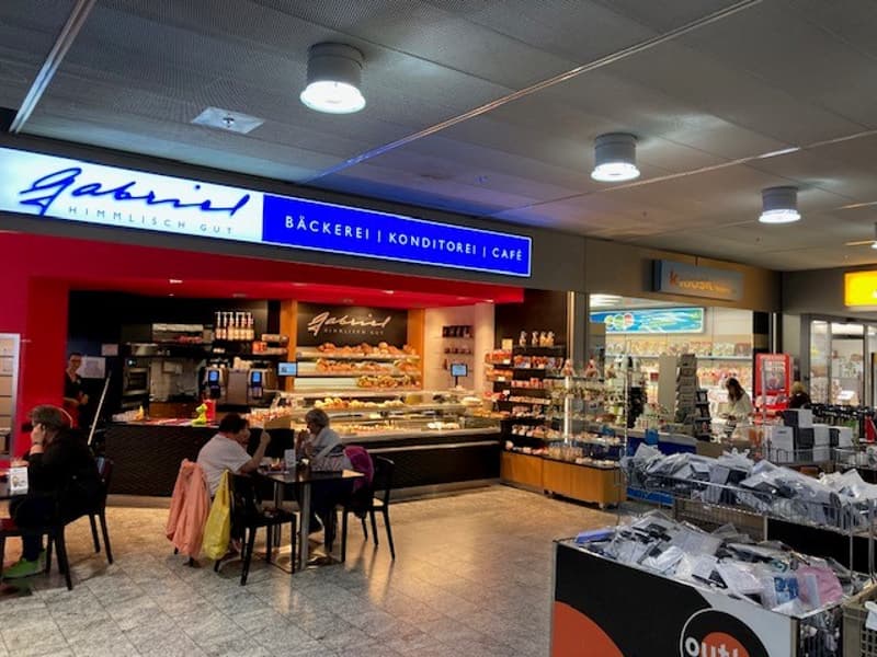 People sitting at tables, counter with food, bakery sign, shelves with items, lights
