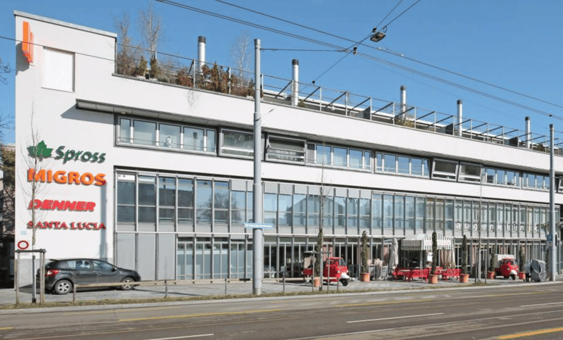 Modern white building with glass windows, multiple floors, signage for Migros and Santa Lucia, outdoor seating area with tables and chairs, small trees, and parked vehicles in front.