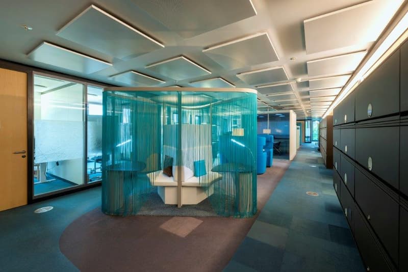 An office space with white walls, blue flooring, ceiling-mounted lamps, a white lounge bench enclosed by a blue curtain, cubicles, and file cabinets. A door leads to another office area.
