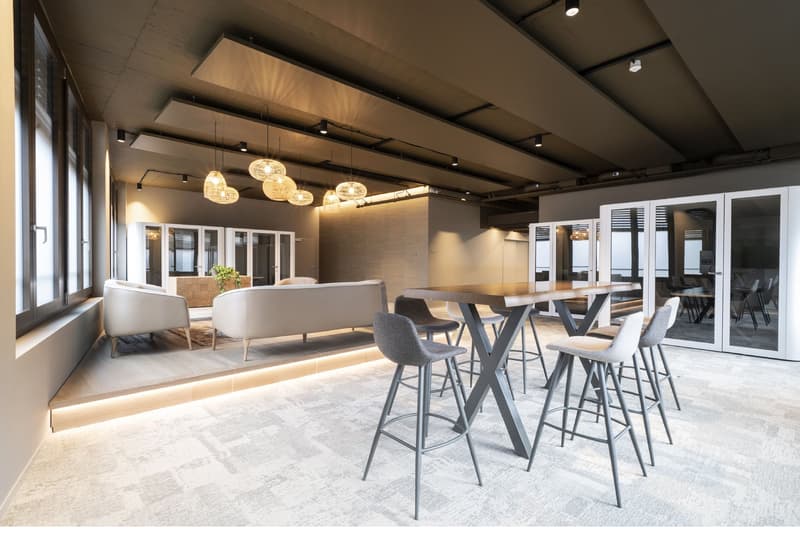 a room with white walls, carpet, two white couches, grey bar stools, a table, a plant, and ceiling lamps