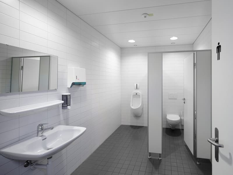 Clean, simple bathroom with white tiled walls, white sink, mirror, urinal, toilet, and gray tiled floor.