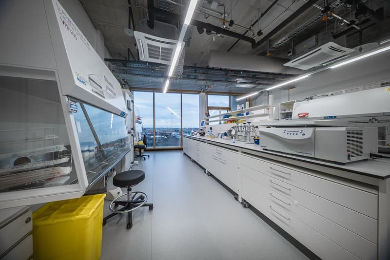White walls, ceiling with equipment, large windows, glass cabinet, yellow bin, several white cabinets, white chairs, white floors, lab equipment.