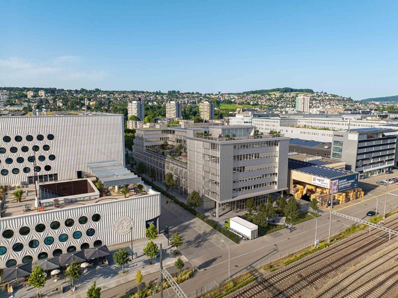 An aerial view of a city with several buildings, one with a circular design, a large building with multiple floors, and a small building with a roof-top garden.
