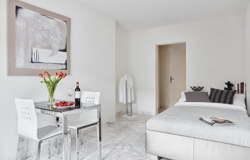 A simple bedroom with a white bed, pillows, and a blanket. A small table with flowers and wine bottle, and chairs. There is a frame on the wall and a white towel rack with a white towel.