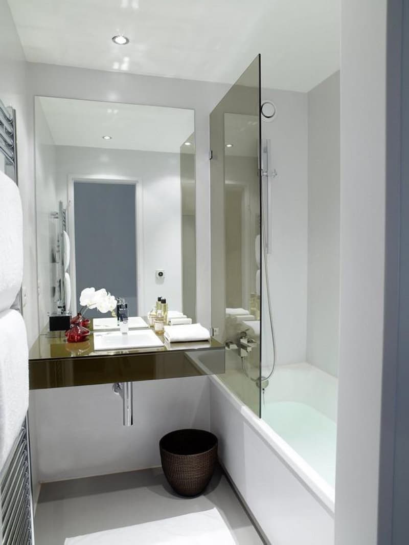 Modern bathroom with white walls, a glass shower, a bathtub, a countertop with a sink, a mirror, and a towel rack. Features include glass paneling and a minimalist design.