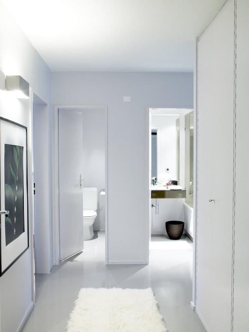 Entrance hallway with white tile flooring, white walls, a rug, and an open door leading to a bathroom with a toilet, sink, and mirror.