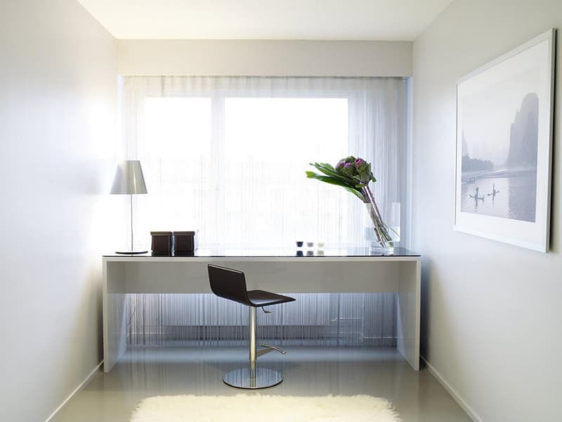 Minimalist modern office with white desk, black swivel chair, desk lamp, window with white curtains, white rug, a framed photograph on the wall.