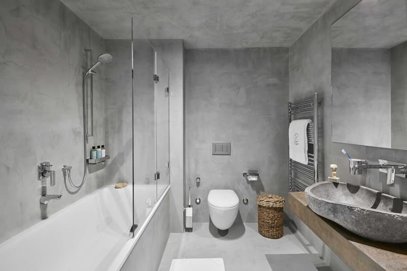 Bathtub, shower, sink, toilet, toilet paper, towels, shaving brush, mirror, grey walls, natural light, small wicker basket, silver toilet paper holder, soap dish