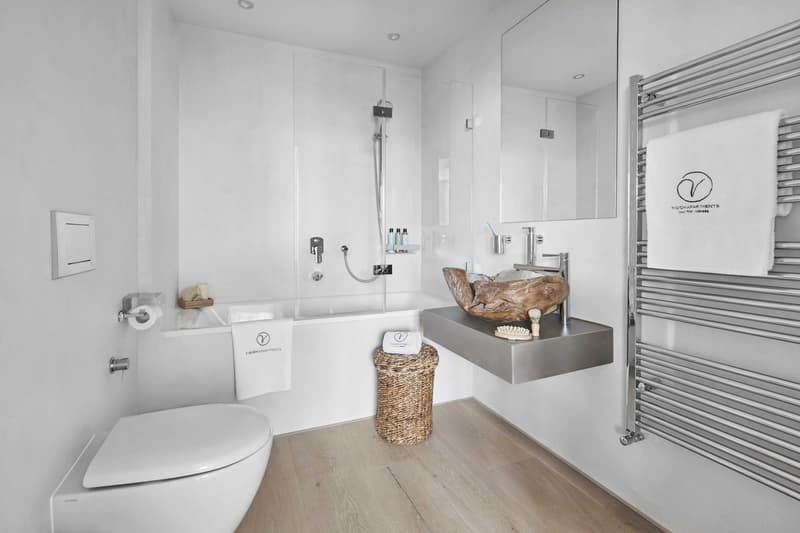 modern white bathroom with a glass walled shower, toilet, large mirror, silver sink, towels, wooden floor, towels rack