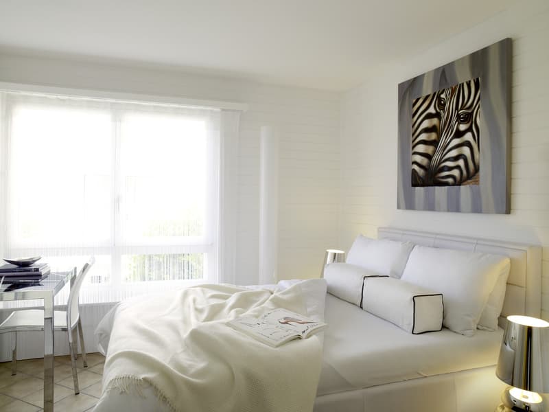 White walls, zebra painting, white bed with white bedding, bedside table with lamp, desk with stack of books, chair, large window with curtains, tiled floor