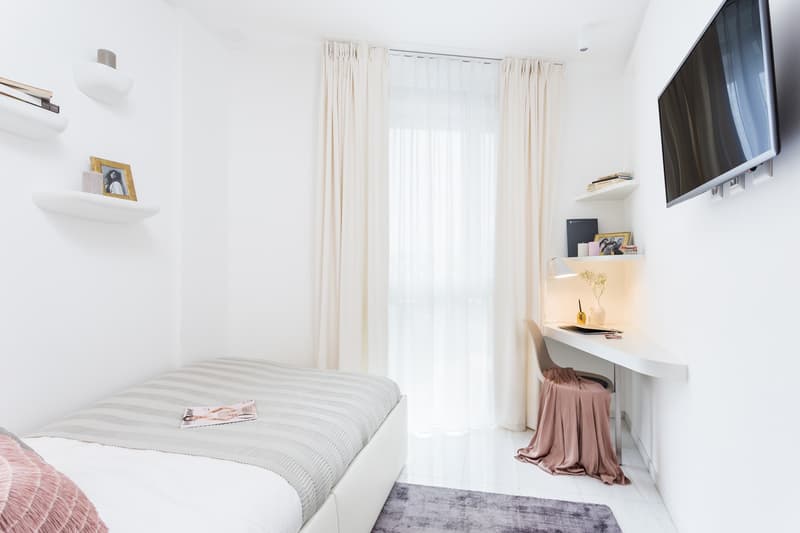 small room with single bed, desk, white shelves, picture frames, tv, large white curtains, pink chair cloth