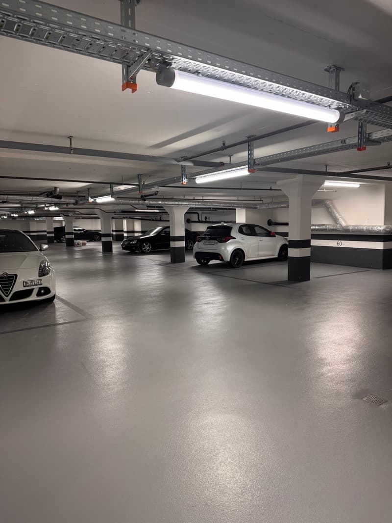 underground parking lot with 3 cars parked, white walls, metal beams, fluorescent lights