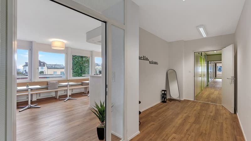 Open space with wooden floor, white walls, large windows, dining area with tables and benches, potted plants, hallway leading to another room, mirror, and open door.