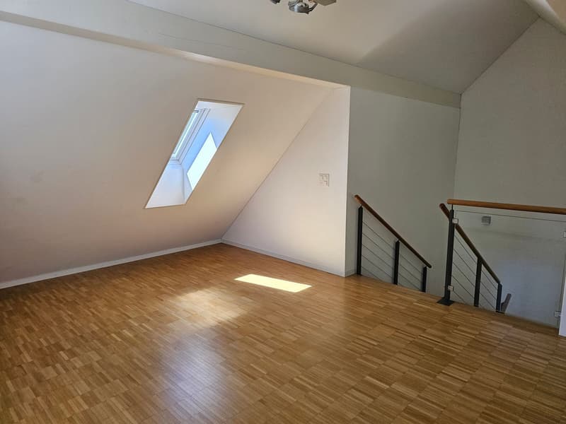 An empty room with wooden flooring and white walls. There is a window on the sloped ceiling and a staircase with a glass railing.