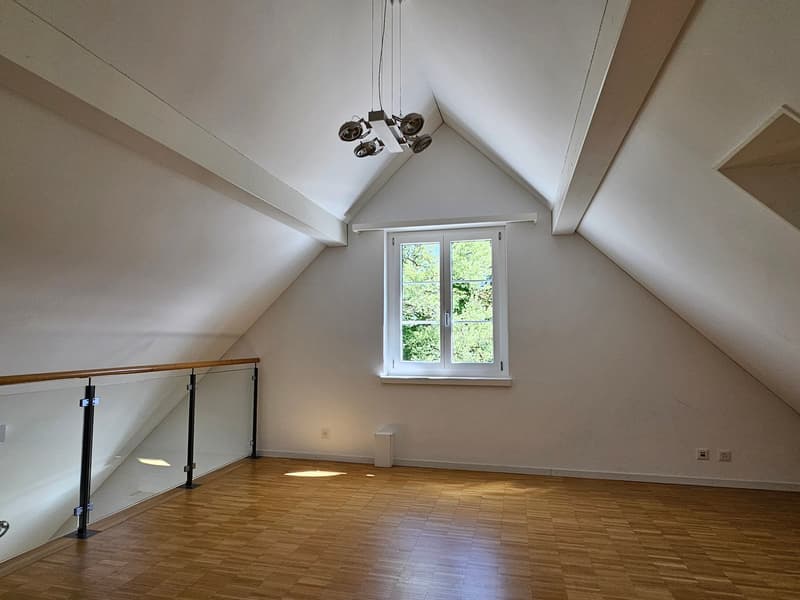 An empty attic room with wooden floors, white walls, a glass railing, and a window with a view of trees outside.