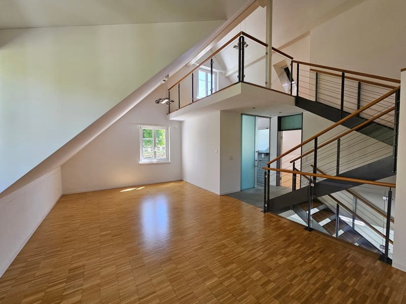 An empty attic space with wooden flooring, white walls, and a glass railing on the upper level. There are two windows, one on the upper level and one on the lower level, and a staircase with a glass railing leading to the lower level.