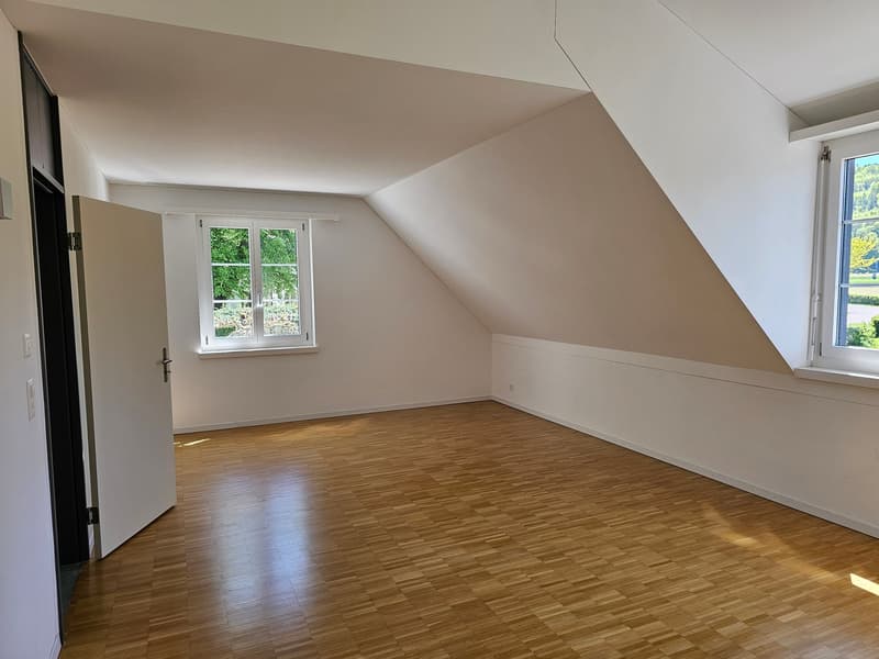 An empty room with wooden flooring, white walls, and two windows. The room has an open door on the left and a slanted ceiling.