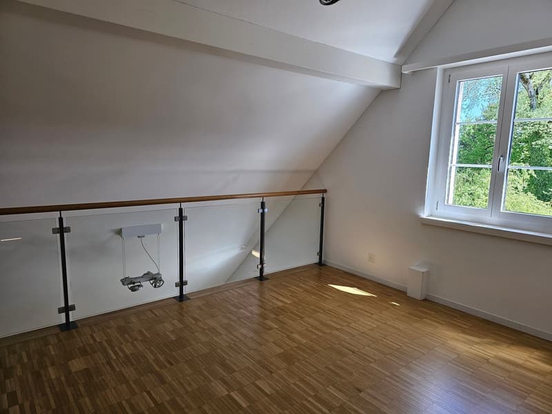 Empty room with wooden flooring, white walls, glass railing, and a window with a view of trees outside.