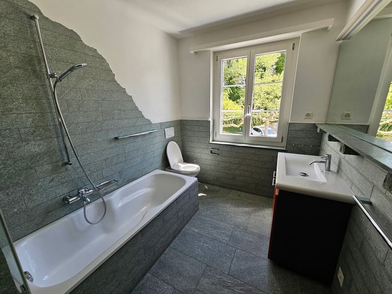 Bathroom with a bathtub, shower, toilet, and sink. The walls and floor are tiled in gray. There is a window with a view of trees outside.