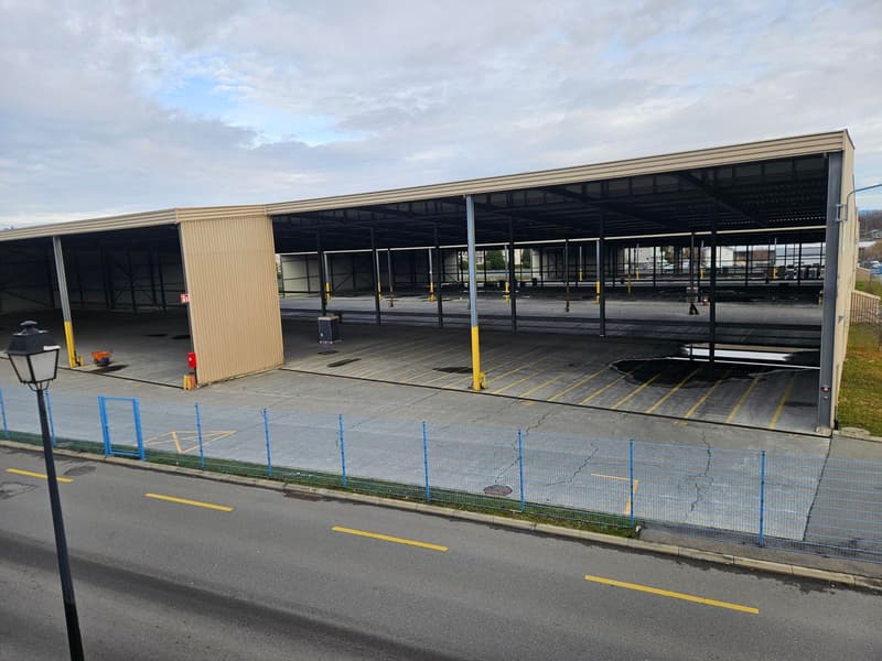 Empty large warehouse, metal roof and walls, blue barriers along the building, yellow lane markings in the parking lot, grey concrete floor, street light, and empty lot behind