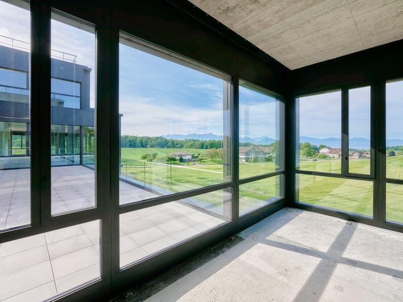 Large windows with a panoramic view of the surrounding countryside, including green fields, trees, and distant mountains. The room has a concrete ceiling and tile flooring.