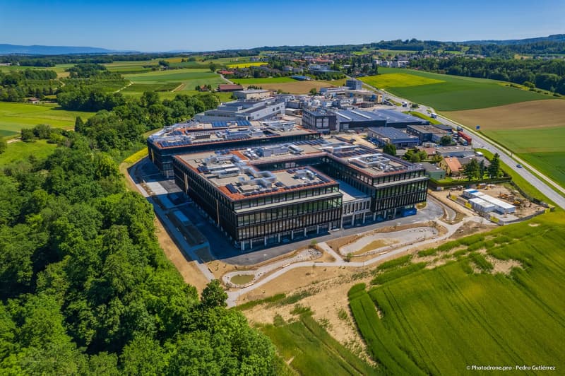 Office building with multiple floors and solar panels, surrounded by greenery and fields.