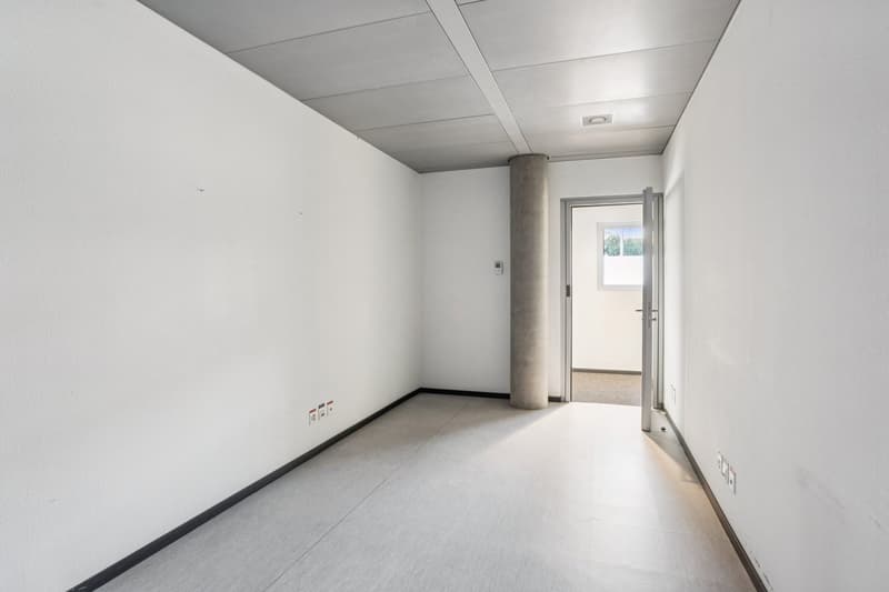 Empty room with white walls, gray floor, central column, and an open door to another room with a window