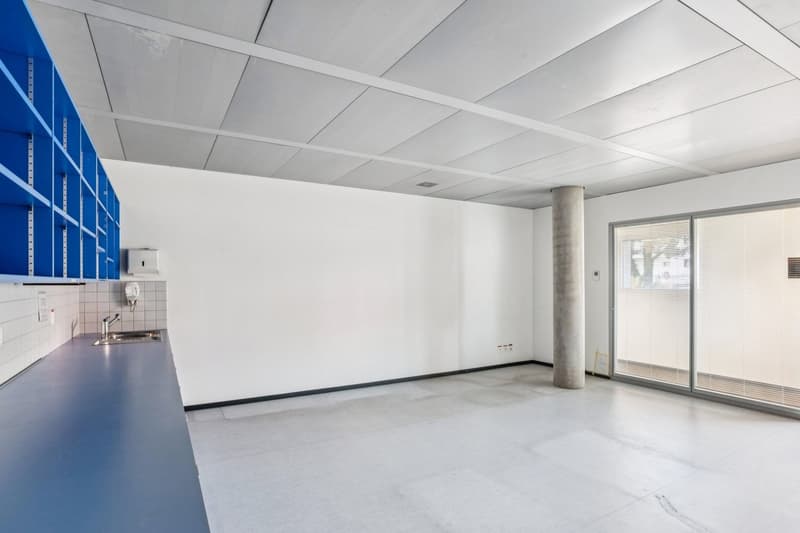 Empty room with white walls, ceiling, and floor. It has a blue cabinet with shelves, a sink with faucet, a power outlet, and a large window with blinds.