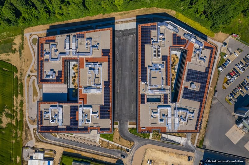 Two building blocks, solar panels on the roof, asphalt driveway, green areas and parking lots with multiple cars