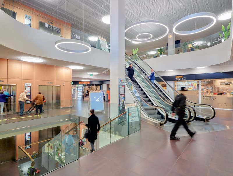 Large indoor shopping mall with glass walls, escalators, and modern lighting fixtures. The mall has various stores and a bistro restaurant visible.