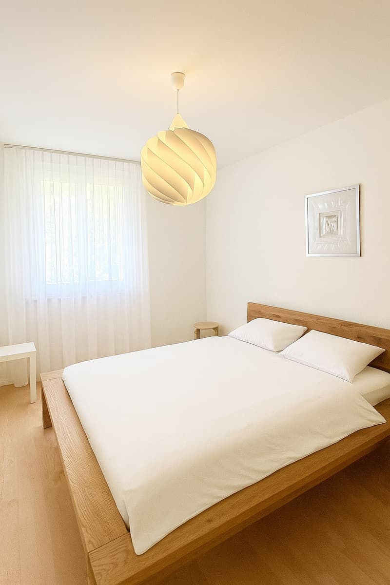 Spacious bedroom with wooden bed, two white pillows, white sheets, wooden nightstand, decorative wall frame, white curtain.