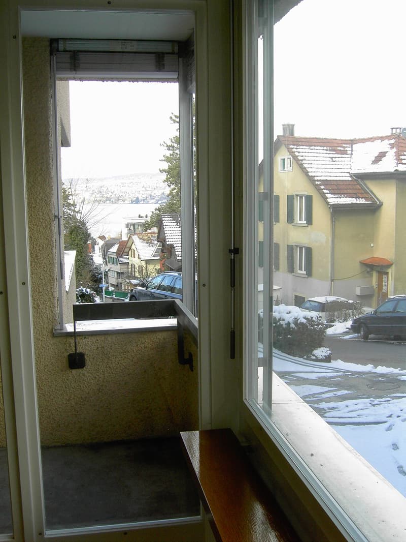 An apartment room with sliding doors to a balcony, carpeted floor, large windows with a view of the street and the landscape