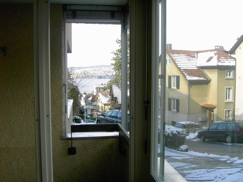 A balcony with a window view, overlooking a snowy street and a house in the distance.