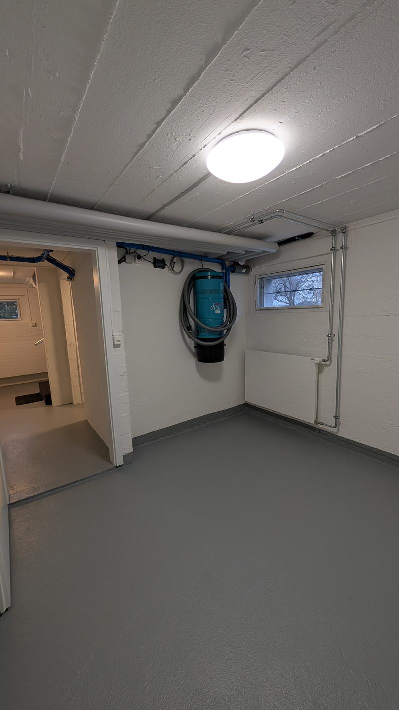Empty room, white walls, gray floor, ceiling light, small window, pipes on the wall, a black object on the wall