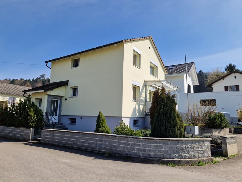 2 story house, yellow exterior, brown roof, white windows, stone wall, plants, paved driveway