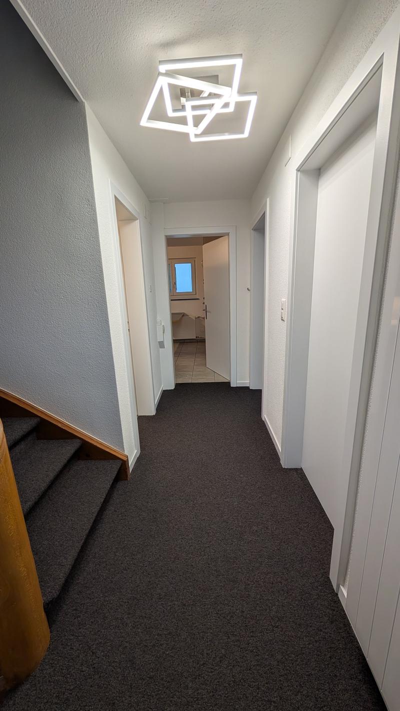 Empty hallway with carpeted floor, white walls, wooden staircase, modern ceiling light, open door leading to a room with a window and a desk.