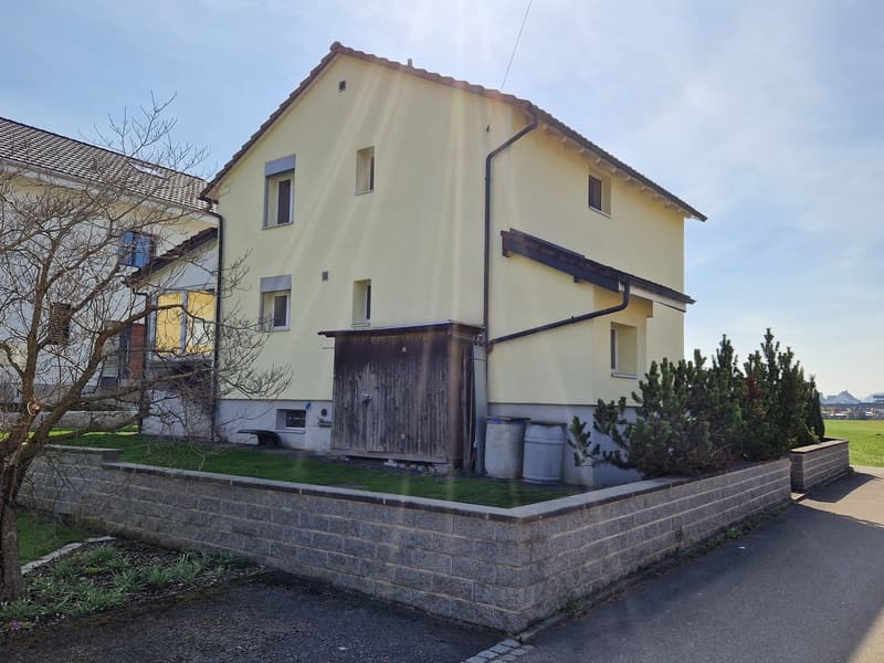Two-story house with yellow exterior walls, brown roof, multiple windows, wooden door, small garden with grass and plants, stone wall, and a driveway.