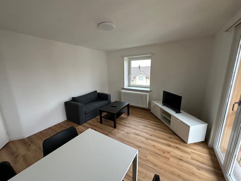 Spacious living room with hardwood flooring, white walls, a black sofa, a coffee table, and a TV stand with a flat-screen TV.